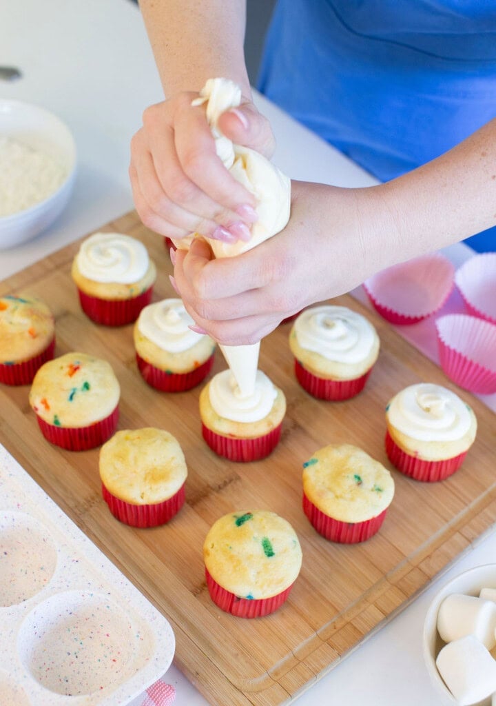 A person is piping white frosting onto cupcakes with colorful sprinkles, each in a red paper liner, on a wooden board. Nearby are empty cupcake liners, a bowl of frosting, and a cupcake tray.