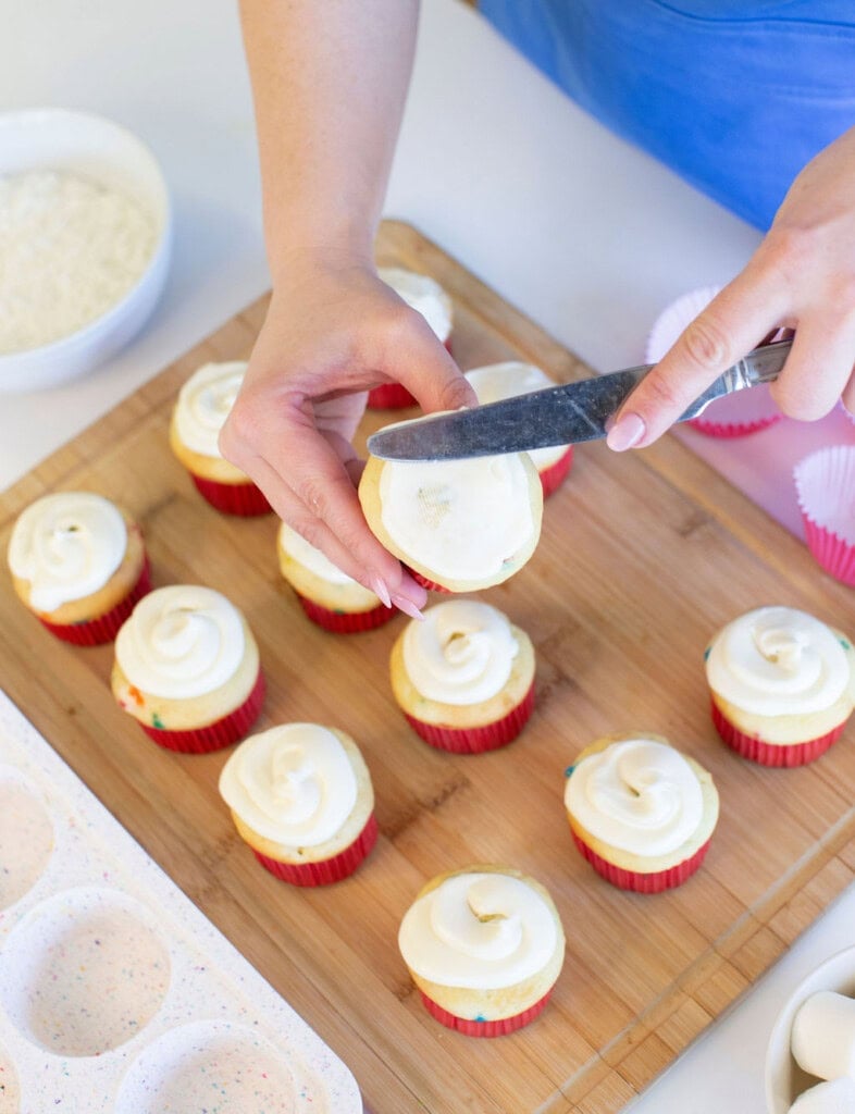 A person spreads white frosting on a cupcake with a knife. Several cupcakes in red liners, already frosted, sit on a wooden board. A bowl of shredded coconut and empty cupcake liners are nearby.