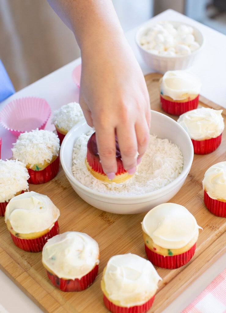 A hand dips a frosted cupcake into a bowl of shredded coconut on a wooden board, surrounded by more cupcakes with white frosting and empty pink cupcake liners.