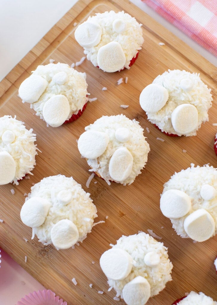 Nine cupcakes decorated with white frosting, shredded coconut, and marshmallows arranged to look like bunny tails and feet, are displayed on a wooden board. A pink checkered cloth is partially visible in the corner.