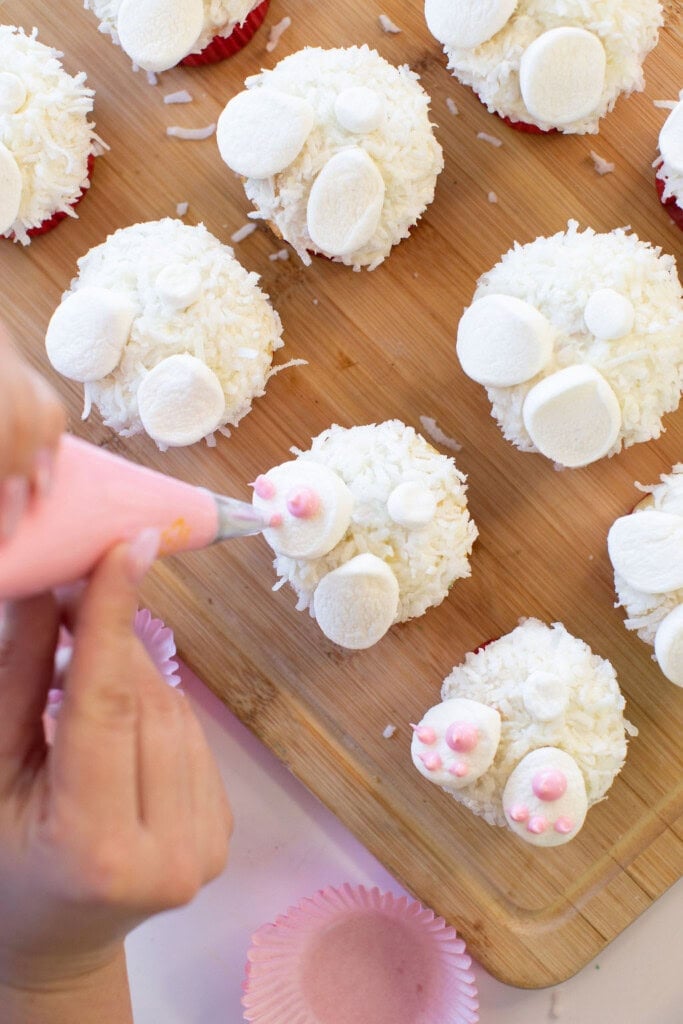 A person decorates cupcakes with pink frosting, creating bunny paw designs using mini marshmallows and shredded coconut. The cupcakes are arranged on a wooden board.