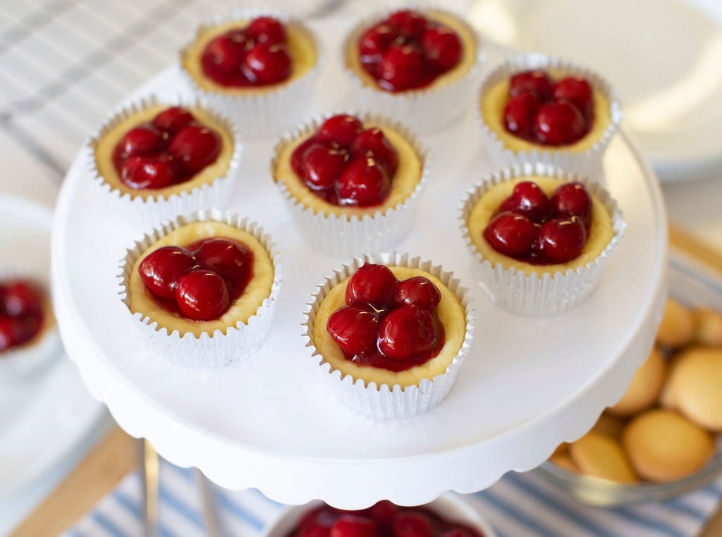 Mini cheesecakes in white cupcake liners topped with glossy red cherries are arranged on a white cake stand. The background includes plates and a tray of small round cookies.