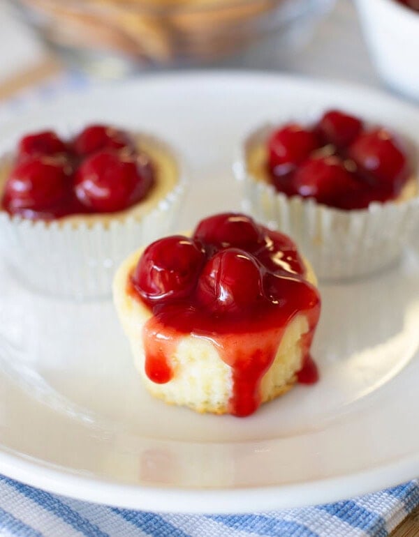 Three mini cheesecakes topped with cherry sauce sit on a white plate. The cheesecakes are in paper liners, and the plate rests on a blue and white striped cloth.