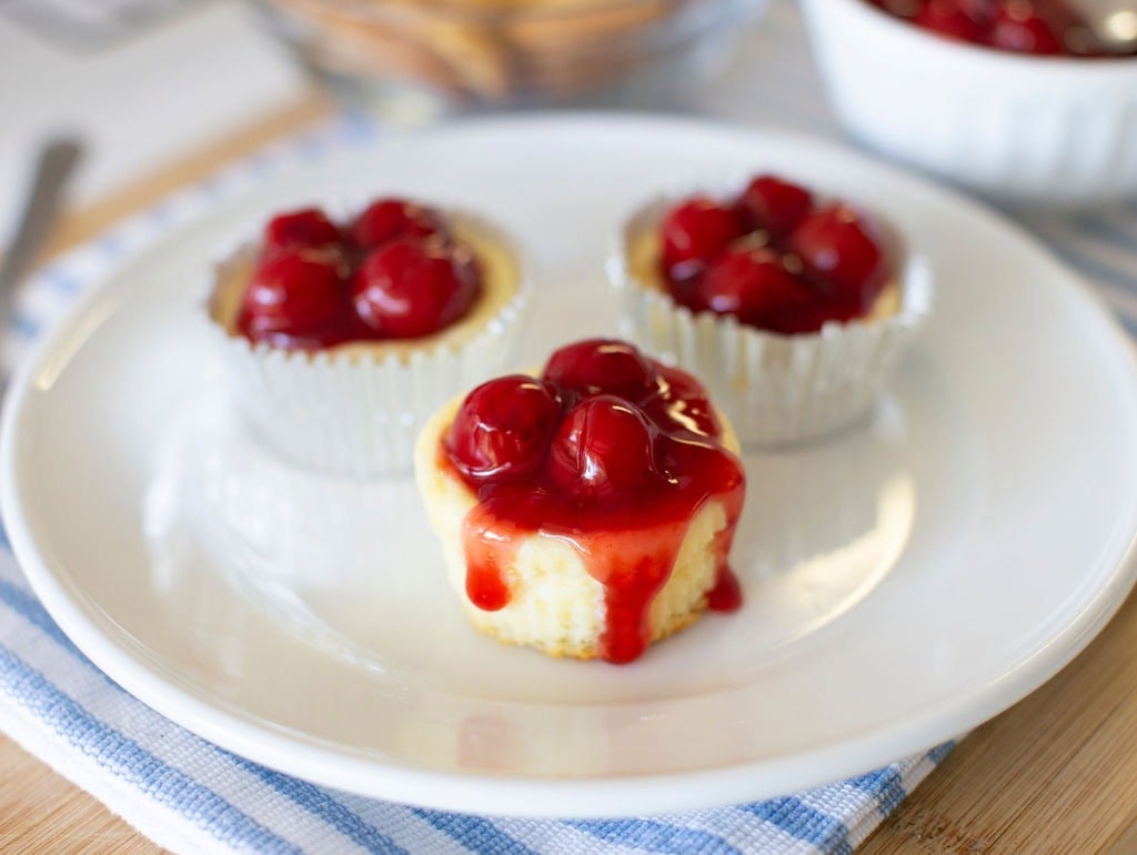 Three mini cheesecakes topped with cherry sauce sit on a white plate. The cheesecakes are in paper liners, and the plate rests on a blue and white striped cloth.