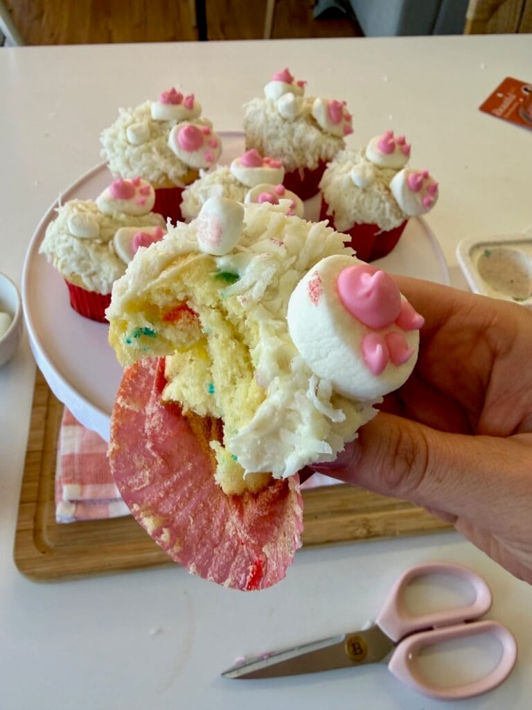 A hand holds a bitten Easter bunny cupcake with white frosting and pink decorations, next to a plate of similarly decorated Easter cupcakes. Pink-handled scissors and a wooden board are on the table nearby.