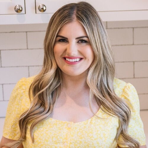 A woman with long, wavy blonde hair and a bright smile stands in a kitchen. She is wearing a yellow floral dress with puffed sleeves and white buttons. The background shows white subway tiles and cabinets.