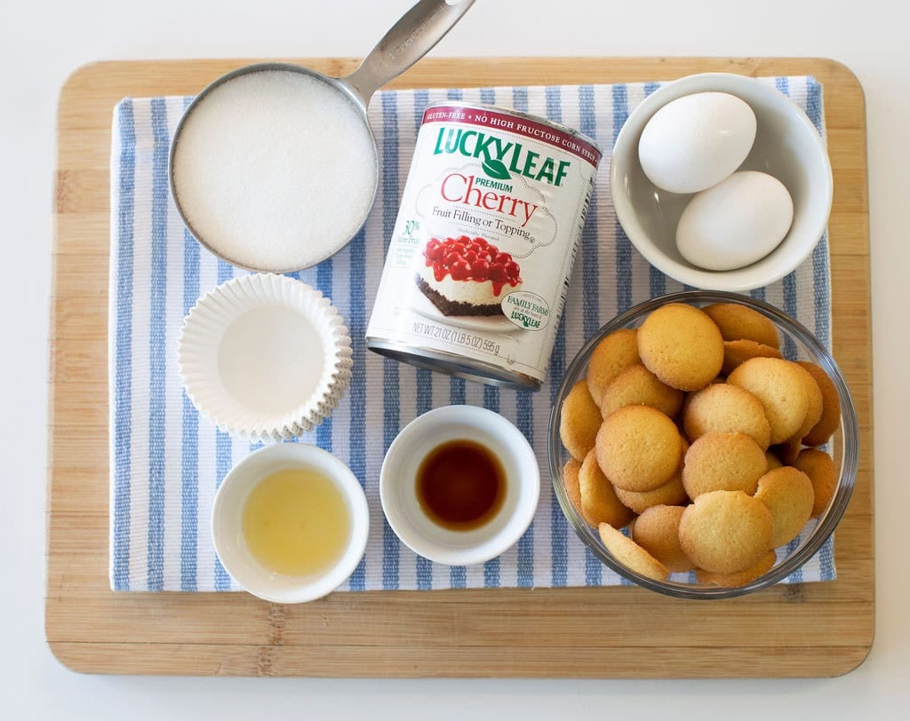 A wooden board with a blue-striped towel holds a measuring cup of sugar, a can of cherry pie filling, three eggs, vanilla wafers in a bowl, cupcake liners, vanilla extract, and lemon juice.