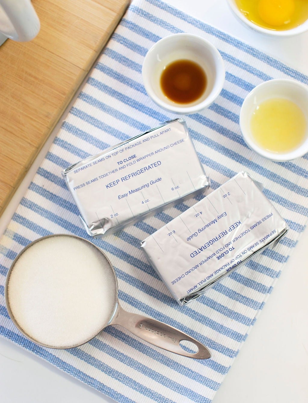 Two blocks of cream cheese in foil, a metal measuring cup filled with sugar, and small bowls containing vanilla extract and egg yolk, all arranged on a blue and white striped cloth.