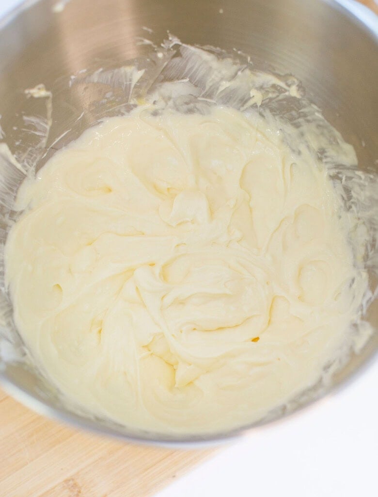 A metal mixing bowl filled with creamy, smooth, pale yellow batter or frosting, sitting on a light wooden surface.