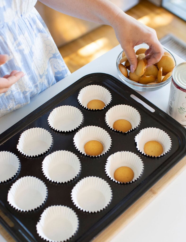 A person places vanilla wafer cookies into paper cupcake liners arranged on a baking tray, with a bowl of cookies and a can nearby on the counter.