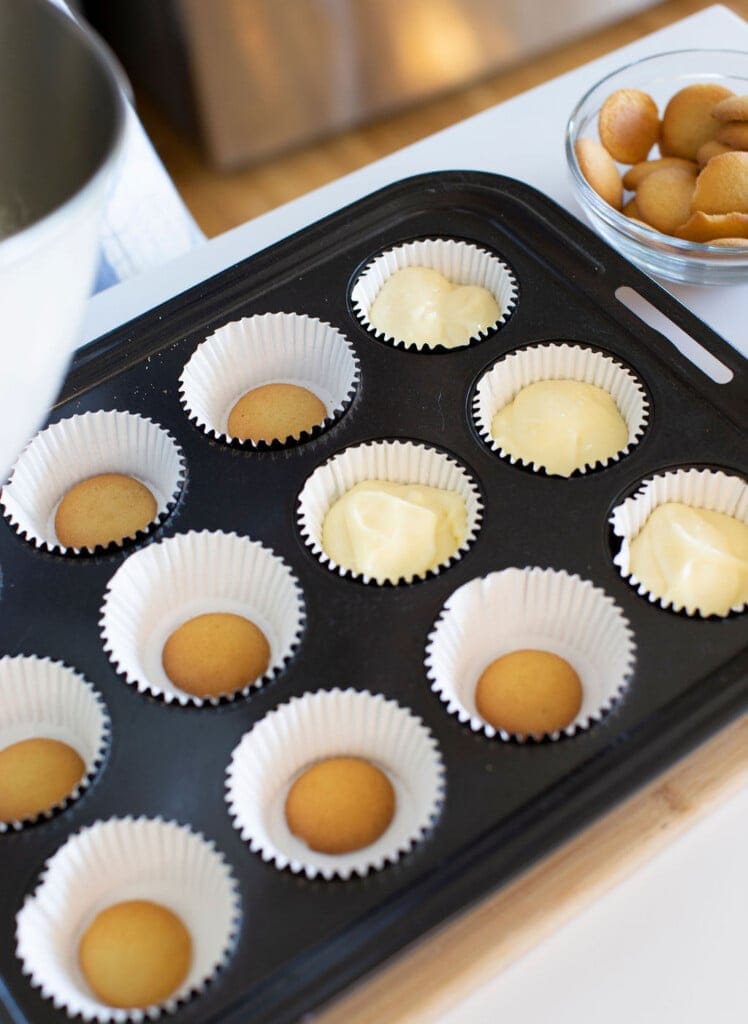 A muffin tin with paper liners, some filled with a cookie at the bottom, others with cookie and creamy batter. A glass bowl of cookies sits nearby on the counter.