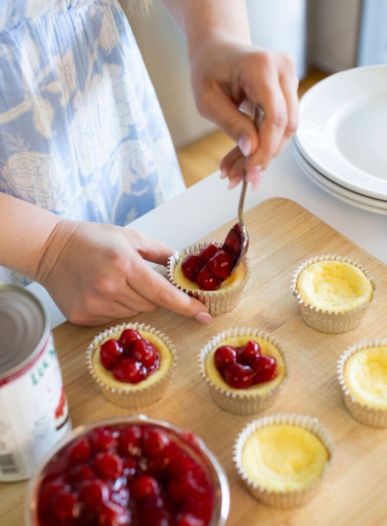 A person in a light blue floral dress is spooning cherry topping onto mini cheesecakes arranged on a wooden cutting board next to a can of cherry filling and a stack of white plates.