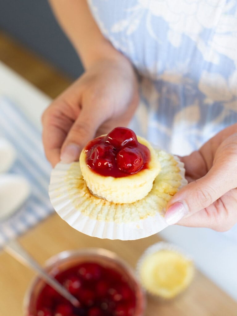 A person holds a mini cheesecake topped with cherry pie filling in a white cupcake liner. A bowl of cherry topping and more mini cheesecakes are on the table below.
