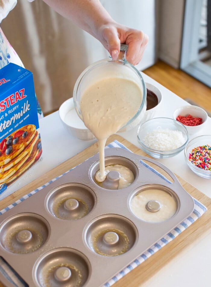 A hand pours pancake batter into a donut-shaped baking pan to make pancake donuts, with bowls of toppings including sprinkles, coconut, and red crumbs nearby, along with a box of Krusteaz Buttermilk Pancake Mix.