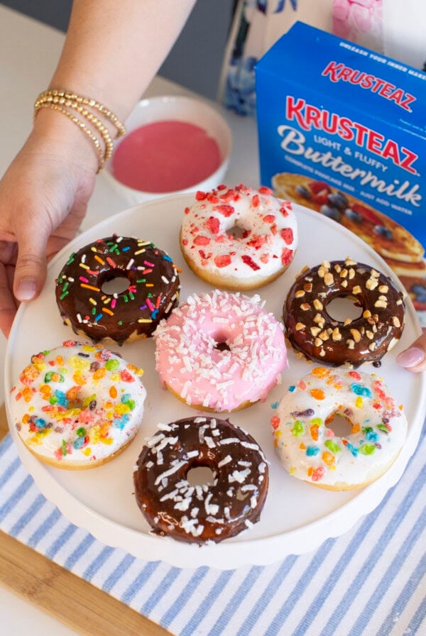 A person holds a white cake stand with eight decorated baked donuts covered in colorful icing, sprinkles, and toppings. A box of Krusteaz Buttermilk Pancake Mix and a bowl of pink icing sit nearby.