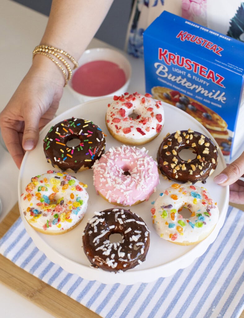 A person holds a white cake stand with eight decorated baked donuts covered in colorful icing, sprinkles, and toppings. A box of Krusteaz Buttermilk Pancake Mix and a bowl of pink icing sit nearby.
