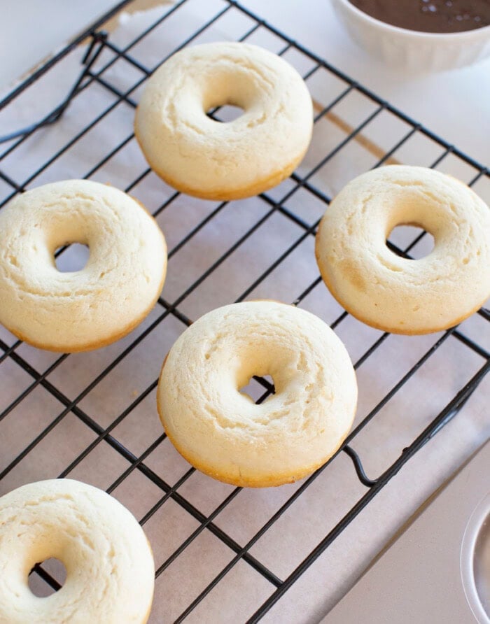Five baked donuts cool on a black wire rack over parchment paper, with a bowl of chocolate glaze in the background.