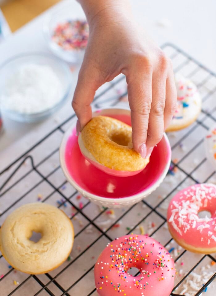 A hand dips a baked donut into pink icing over a wire rack with decorated donuts and bowls of sprinkles and powdered sugar in the background.