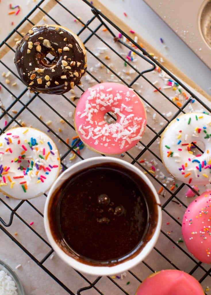 A cooling rack holds baked donuts with pink, white, and chocolate glaze, topped with sprinkles and nuts. A bowl of chocolate glaze sits nearby with colorful sprinkles scattered around.