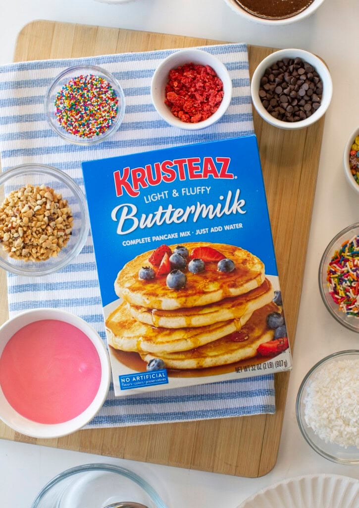 A box of Krusteaz Buttermilk Pancake Mix sits on a striped cloth with bowls of colorful sprinkles, nuts, pink icing, chocolate chips, and other toppings nearby for decorating baked donuts.