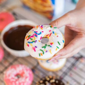 A hand holds a pancake donut topped with white icing and colorful sprinkles above a cooling rack with more decorated pancake donuts and a bowl of chocolate glaze in the background.