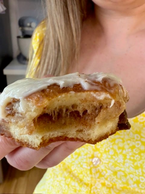 A woman in a yellow floral dress holds up a cinnamon roll with cream cheese icing, showing a large bite taken out of it. The soft, gooey interior and cinnamon filling are visible.