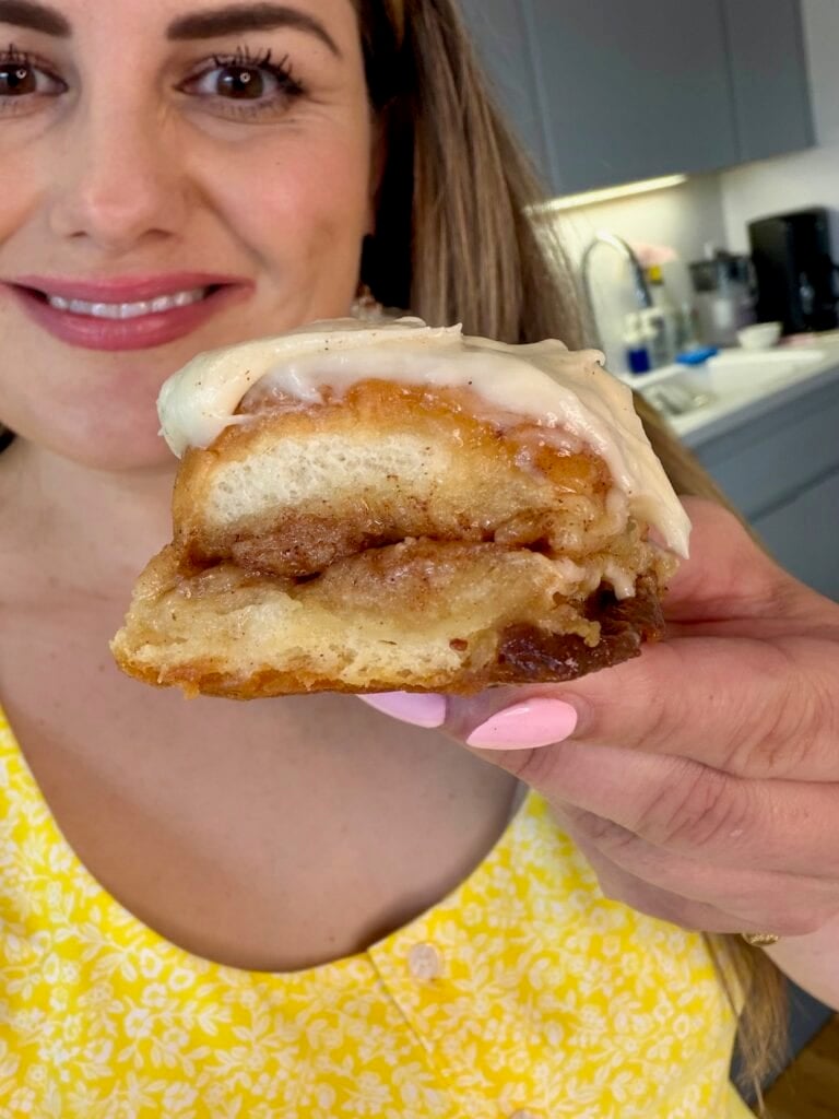 A woman in a yellow dress holds up a gooey cinnamon roll topped with icing, smiling in a kitchen setting. The focus is on the cinnamon roll, showing its swirled layers and creamy frosting.