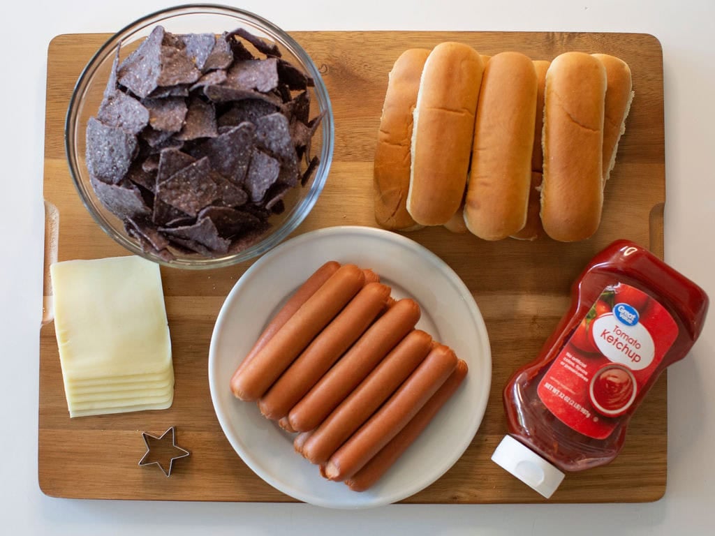 A wooden board holds a bowl of blue corn chips, three hot dog buns, a plate with six hot dogs, slices of cheese, a bottle of ketchup, and a small star-shaped cookie cutter.