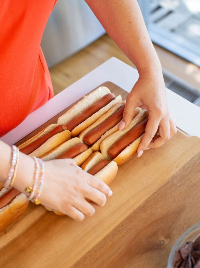 A person in an orange shirt arranges several hot dogs in buns on a wooden board, preparing food. Only the persons arms and hands are visible.