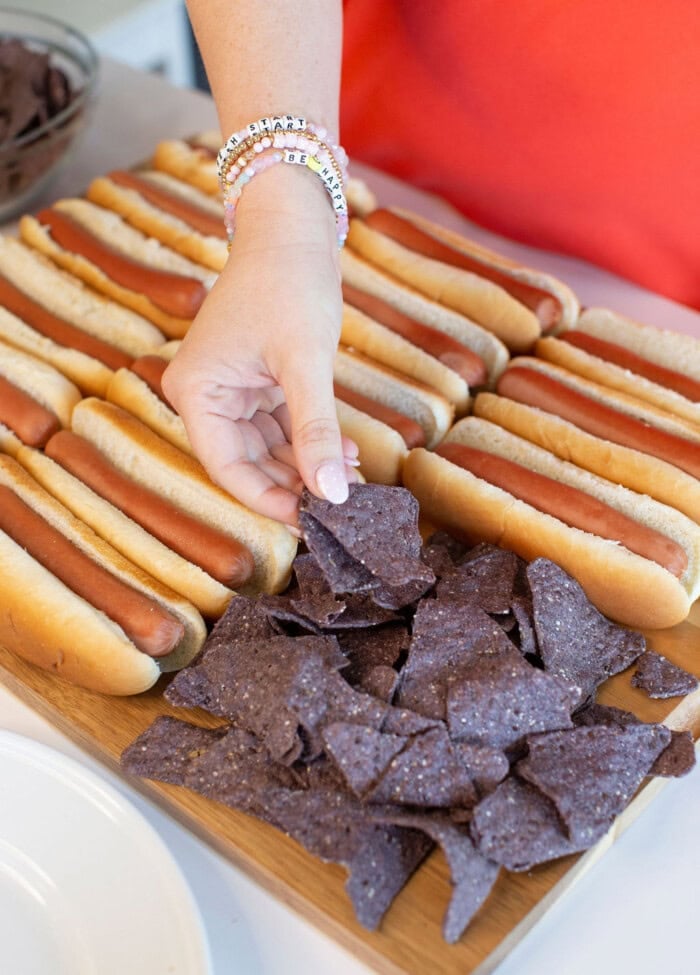 A person wearing beaded bracelets grabs blue corn tortilla chips from a wooden board next to several hot dogs in buns.
