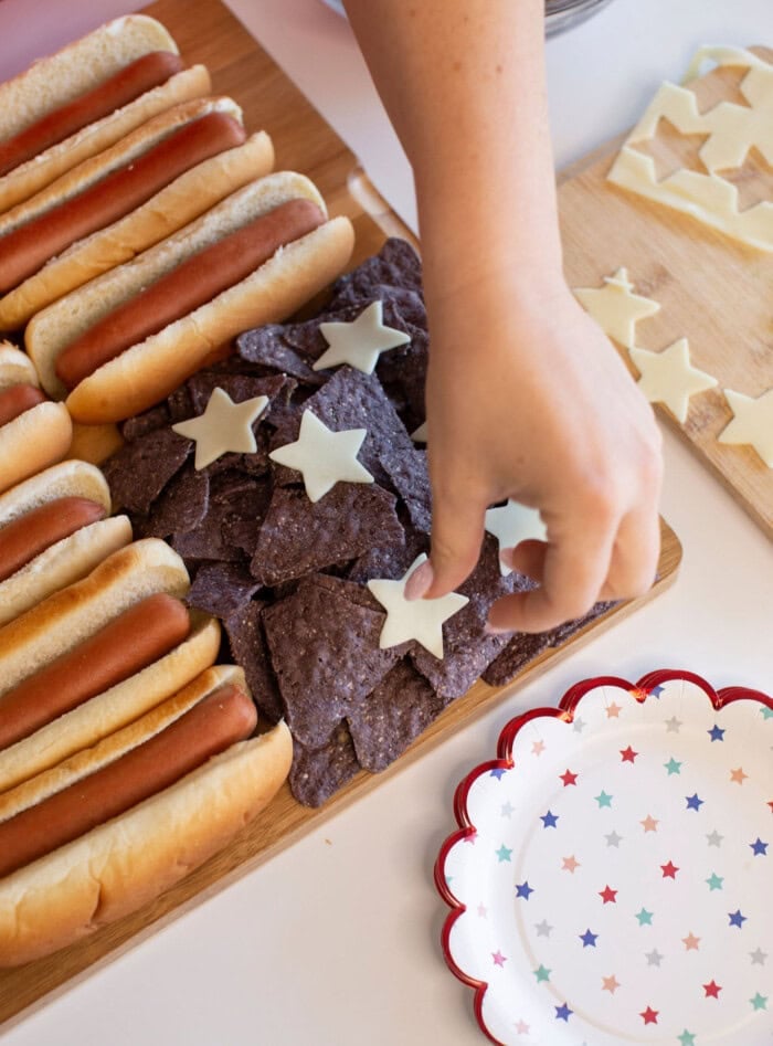 A hand places star-shaped cheese slices onto blue corn tortilla chips beside several hot dogs on a wooden board. Star-shaped cheese cutouts and a star-patterned plate are also visible.