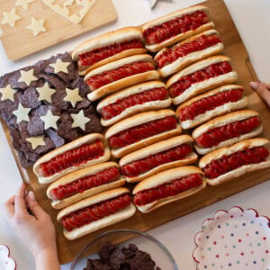 A wooden board displays hot dogs with ketchup arranged in rows and blue tortilla chips with cheese stars, resembling the American flag. Hands are holding the board, and star-patterned plates surround it.