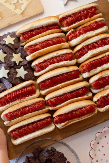 A wooden board displays hot dogs with ketchup arranged in rows and blue tortilla chips with cheese stars, resembling the American flag. Hands are holding the board, and star-patterned plates surround it.