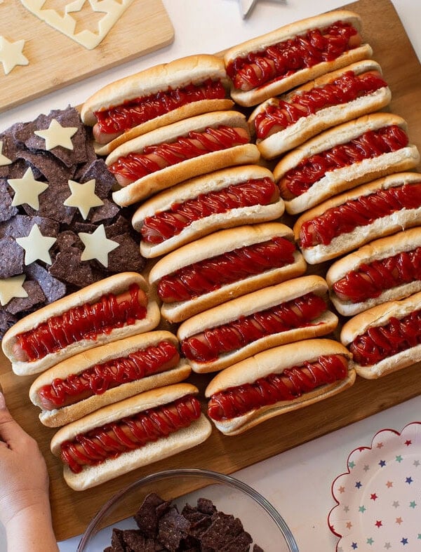 A wooden board displays hot dogs with ketchup arranged in rows and blue tortilla chips with cheese stars, resembling the American flag. Hands are holding the board, and star-patterned plates surround it.