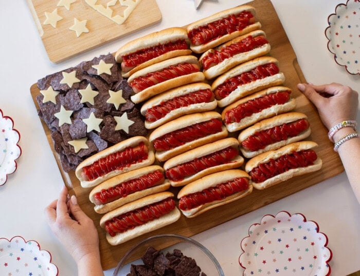A wooden board displays hot dogs with ketchup arranged in rows and blue tortilla chips with cheese stars, resembling the American flag. Hands are holding the board, and star-patterned plates surround it.