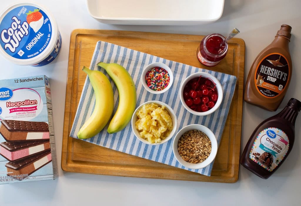 A wooden tray holds two bananas, bowls of pineapple, cherries, nuts, sprinkles, and a jar of strawberry topping on a striped cloth. Surrounding the tray are ice cream sandwiches, whipped topping, and chocolate syrups.