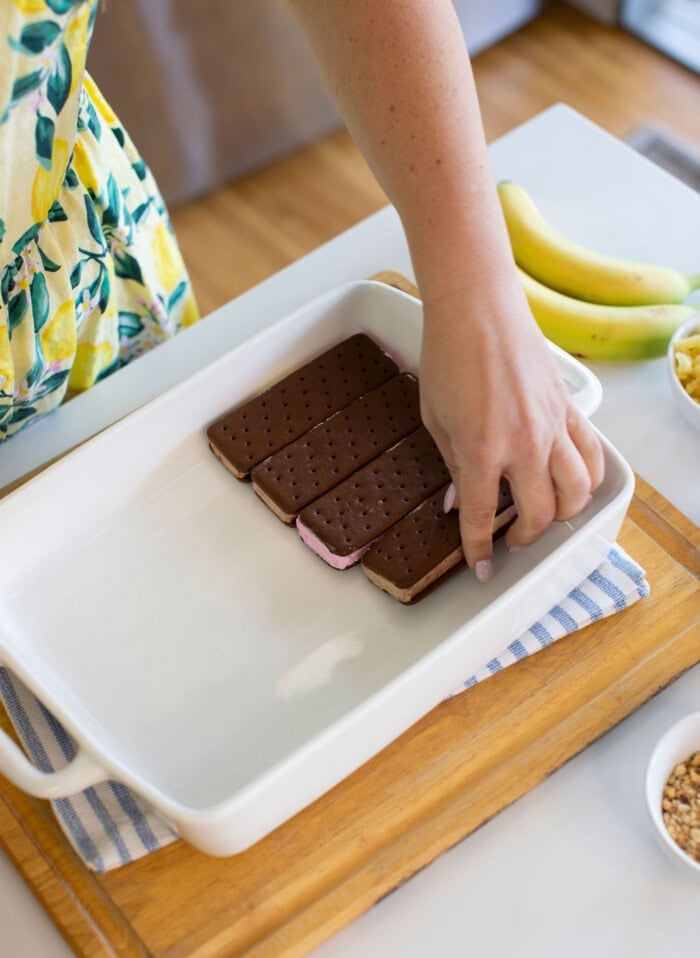 A person places ice cream sandwiches in a white baking dish on a wooden board; bananas and small bowls with toppings are nearby.