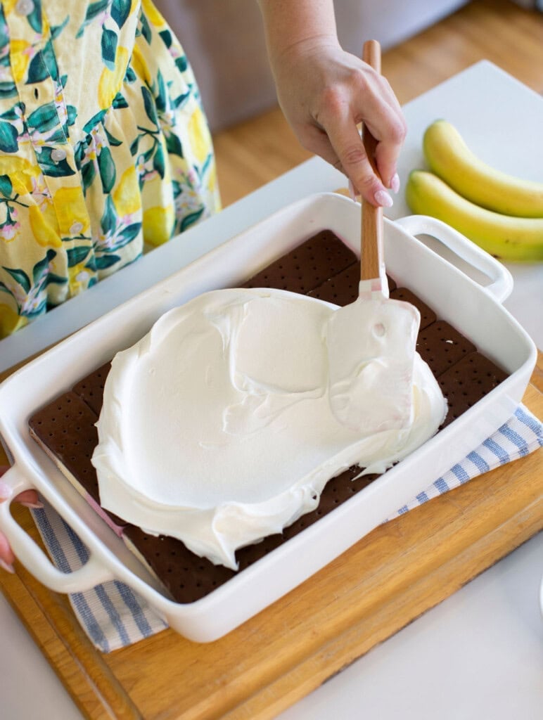 A person spreads a thick layer of whipped topping over ice cream sandwiches in a white baking dish. Two bananas and a blue-striped cloth are on the counter nearby. The person is wearing a yellow, floral-patterned dress.