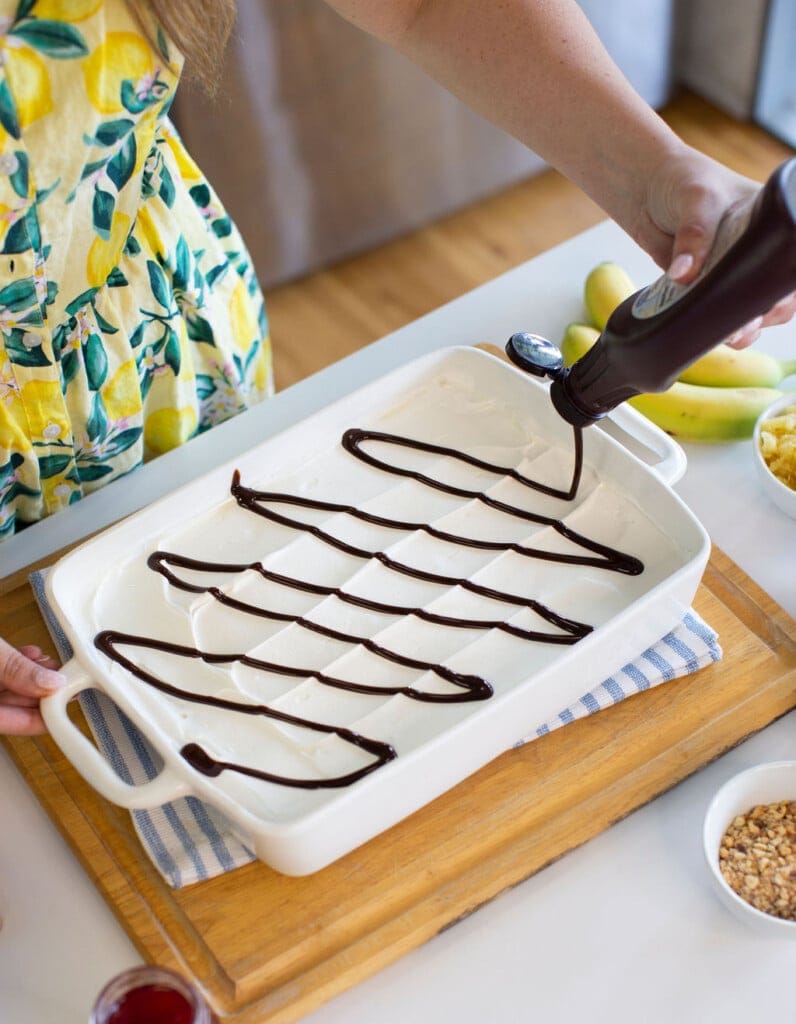 A person in a yellow dress with a lemon pattern is drizzling chocolate syrup in a grid pattern over a layer of vanilla ice cream in a white rectangular dish on a wooden tray. Nearby are bananas and bowls with chopped nuts.