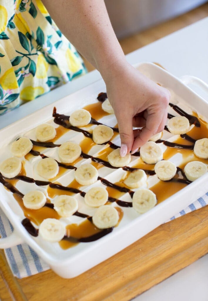A hand places banana slices on a white dish drizzled with caramel and chocolate sauce. The person is wearing a yellow and green dress with a lemon pattern. The dish rests on a blue-striped towel and wooden surface.