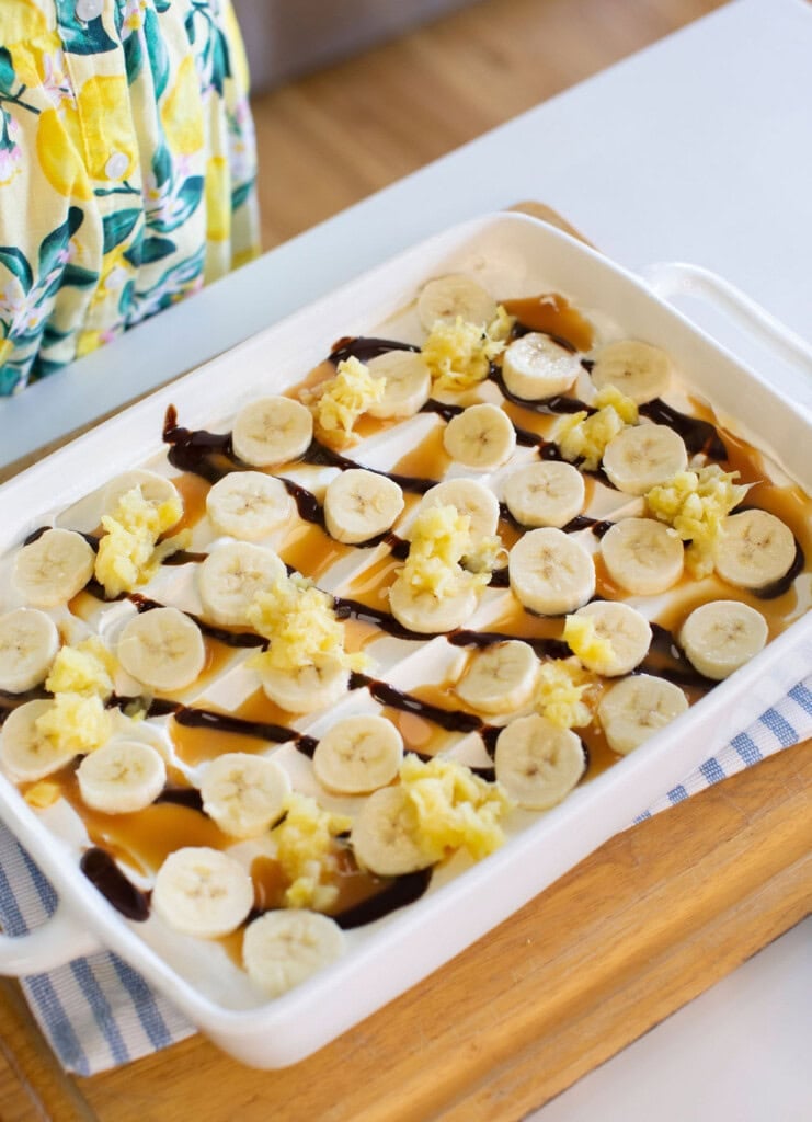 A white baking dish filled with a dessert topped with banana slices, caramel and chocolate drizzle, and small scoops of what appears to be crushed pineapple. A person in a lemon-patterned apron stands nearby.