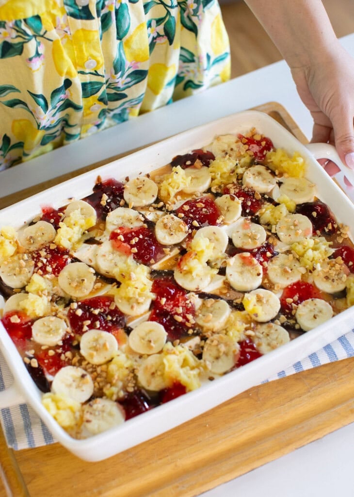 A person in a yellow, lemon-patterned dress holds a white baking dish filled with sliced bananas, dollops of red jelly, crushed nuts, and other toppings, placed on a wooden tray.