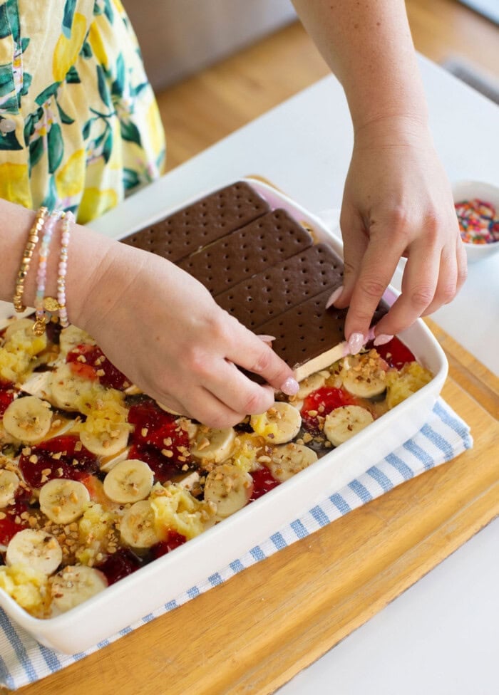 A person places chocolate ice cream sandwiches on top of a dessert layered with banana slices, pineapple, strawberry sauce, and crushed nuts in a white dish. The dish sits on a striped cloth on a wooden tray.