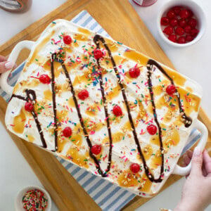 A large ice cream sundae dessert in a white dish, topped with whipped cream, rainbow sprinkles, maraschino cherries, chocolate syrup, and caramel sauce. Surrounding bowls hold cherries, sprinkles, and syrup.