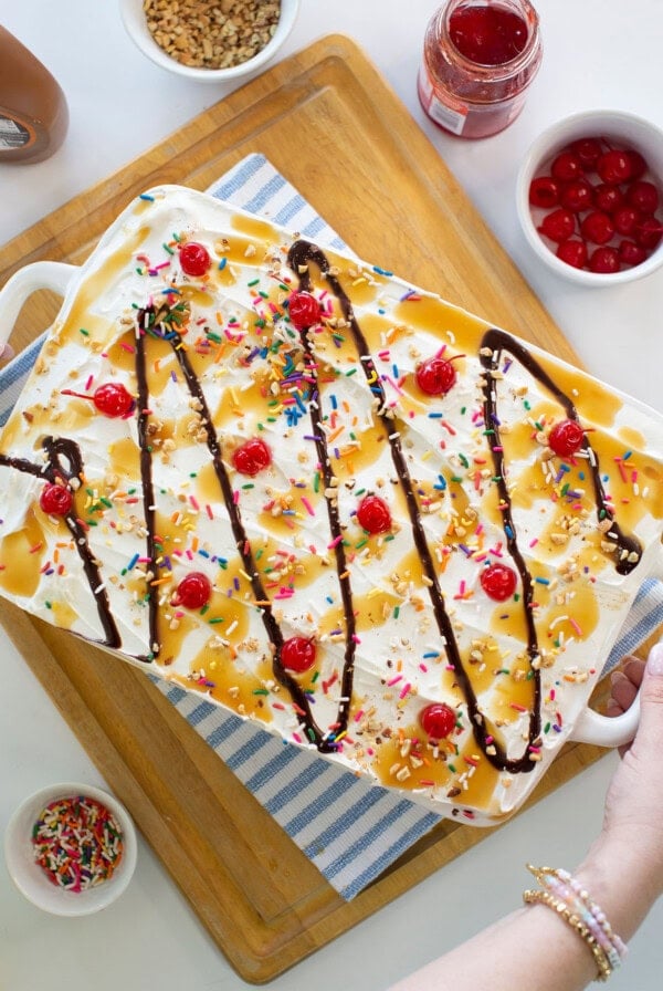A large ice cream sundae dessert in a white dish, topped with whipped cream, rainbow sprinkles, maraschino cherries, chocolate syrup, and caramel sauce. Surrounding bowls hold cherries, sprinkles, and syrup.
