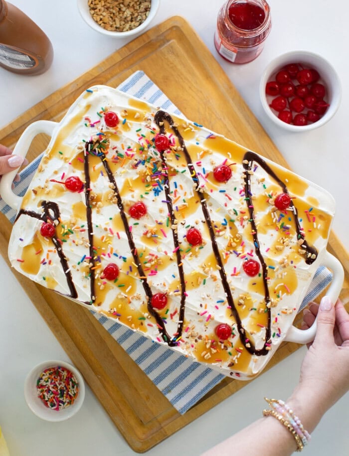 A large ice cream sundae dessert in a white dish, topped with whipped cream, rainbow sprinkles, maraschino cherries, chocolate syrup, and caramel sauce. Surrounding bowls hold cherries, sprinkles, and syrup.