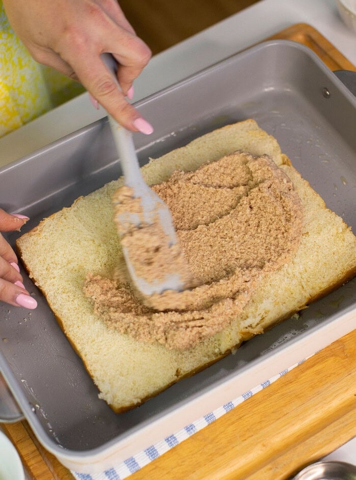A person spreads a brown, crumbly mixture onto a rectangular layer of cake in a metal baking pan using a spatula. The persons hands are visible, and the cake is on a wooden board.