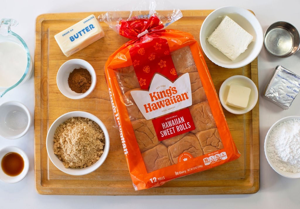 A package of Kings Hawaiian sweet rolls on a wooden board surrounded by small bowls of ingredients, including butter, cream cheese, milk, brown sugar, cinnamon, vanilla extract, and powdered sugar.