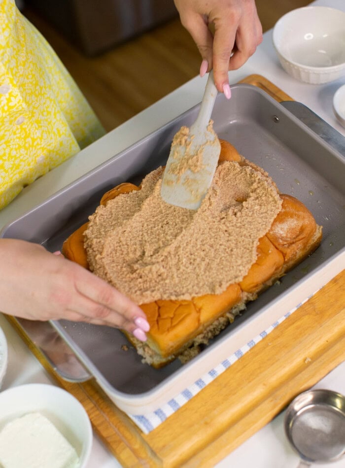 A person spreads a brown, crumbly mixture over a layer of bread rolls in a baking pan using a spatula. Bowls with ingredients and kitchen tools are visible on the counter nearby.