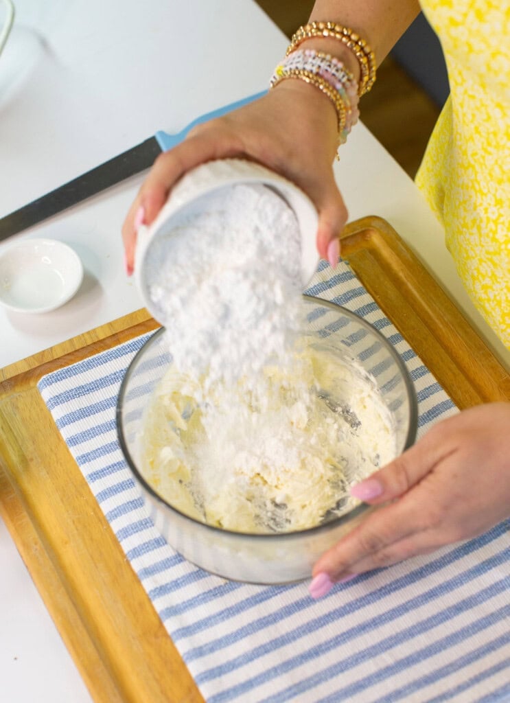 A person pours powdered sugar from a white bowl into a glass mixing bowl containing a creamy mixture, on a wooden tray with a striped cloth underneath.